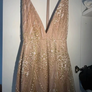 Champagnefärgad glittrig balklänning  - Har aldrig haft på mig denna - pris kan diskuteras. Säljer en champagnefärgad långklänning med djup v-ringning och glittriga paljettdetaljer i guld och vitt. Klänningen har ärmlös design och ett elegant fall. Perfekt för balen!