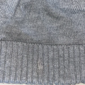 Grå stickad mössa från Polo Ralph Lauren - Snygg grå stickad mössa från Polo Ralph Lauren med diskret broderad logga framtill. Klassisk ribbad kant och mjukt material som håller dig varm under kyliga dagar. Perfekt accessoar för en stilren look.
