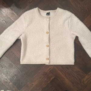 Beige bouclejacka från Gina Tricot - Säljer en beige bouclejacka från Gina Tricot med rund halsringning och fyra dekorativa guldfärgade knappar framtill. Jackan är croppad och har lång ärm, perfekt för att lyfta en enkel outfit.