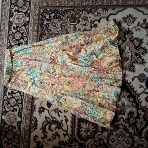 Färgglad blommig långkjol - Säljer en långkjol med blommigt mönster i gula, gröna och orange toner. Kjolen har en luftig och flowy passform med volangdetaljer och resår i midjan. Perfekt för varma dagar och somriga outfits.