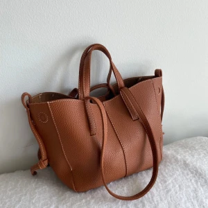 Brun handväska - Säljer en snygg brun handväska med två handtag och en lång axelrem. Väskan har en stilren design med struktur i materialet och är perfekt för att bära dina vardagsprylar. Enkel att matcha till olika outfits!