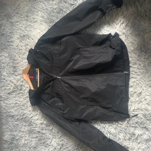 Svart vindjacka från Polo Ralph Lauren - Säljer en svart vindjacka från Polo Ralph Lauren med huva och dragkedja framtill. Jackan har två sidofickor med dragkedja och justerbar nederkant. Perfekt för blåsiga dagar och enkel att matcha med det mesta. Sälj på grund av att den är för stor för mig. 