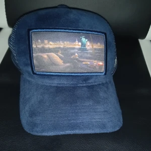 Mörkblå keps med mesh och tryck från Natone - Snygg mörkblå keps med mesh på sidorna och bak, samt ett tryckt motiv framtill. Märket Natone är broderat på sidan. Perfekt accessoar för en cool och avslappnad stil. 