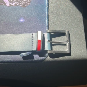 Mörkblått skärp från Tommy Hilfiger - Snyggt mörkblått bälte från Tommy Hilfiger i äkta skinn. Bältet har en klassisk metallspänne och en liten detalj med röd, vit och blå rand vid fästet. Längd 85 cm (34 tum). Perfekt för att lyfta din outfit.