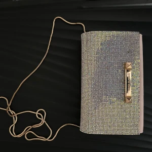Glittrig kuvertväska med kedja - Säljer en glittrig kuvertväska från River Island med guldfärgad kedja och dekorativa detaljer framtill. Perfekt för att lyfta din outfit med extra bling. Väskan är kompakt och smidig att bära över axeln. Du står för frakt, Andvänd 2 gånger