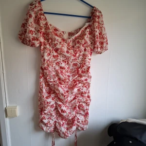 Blommig kortklänning med puffärm - Söt kortklänning med rödblommigt mönster på vit botten. Klänningen har puffärmar, rynkade detaljer framtill och knytband nertill. Perfekt för sommarens alla tillfällen.