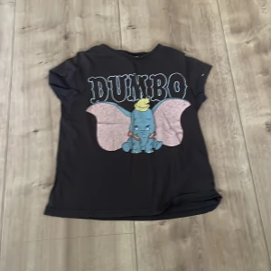 Svart Dumbo t-shirt från Disney - Svart t-shirt från Disney med stort tryck av Dumbo på framsidan. T-shirten har rund hals och korta ärmar. Perfekt för dig som gillar klassiska Disney-karaktärer och vill ha en bekväm och snygg t-shirt.
