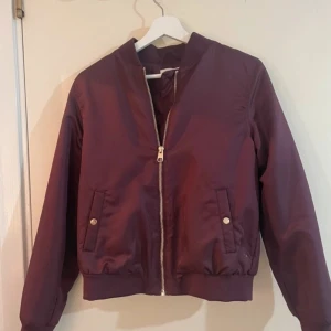 Vinröd bomberjacka med dragkedja - Säljer en vinröd bomberjacka med silvrig dragkedja framtill och ribbade muddar vid ärmslut och midja. Jackan har två fickor med knappstängning och klassisk bomberkrage. Perfekt för dig som vill ha en stilren och enkel jacka.
