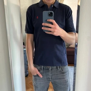 Mörkblå pikétröja från Polo Ralph Lauren - Säljer en klassisk mörkblå pikétröja från Polo Ralph Lauren med röd broderad logga på bröstet. Tröjan har krage och knappar vid halsen, samt en normal passform. Perfekt för en stilren och avslappnad look.