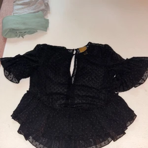 Svart blus med volang och prickigt mönster - Säljer en svart blus i storlek 32 med volang nertill och vid ärmslut. Blusen har ett diskret prickigt mönster i tyget . 