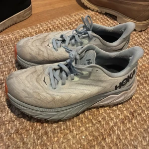 Ljusblå sneakers från Hoka  - Säljer ett par ljusblå sneakers från Hoka. Modell Arachi 6 med tjock sula och snörning. Skorna har en sportig design med diskreta reflexdetaljer och orange detalj vid tån. Perfekta för träning eller vardagsbruk. 