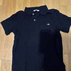 Mörkblå pikétröja från GANT - Klassisk mörkblå pikétröja från GANT med broderad logga på bröstet. Tröjan har korta ärmar, krage och två knappar framtill. Tillverkad i mjuk bomull som känns skön mot huden. Perfekt för en clean och stilren look.