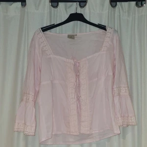 Ljusrosa blus med spets från Vero Moda - Romantisk ljusrosa blus från Vero Moda med vackra spetsdetaljer längs halsringning, ärmslut och framtill. Blusen har trekvartsärmar, knytning framtill och en lätt, luftig känsla. Perfekt för dig som gillar en feminin och bohemisk stil. Kontakta för fler frågor eller bilder.