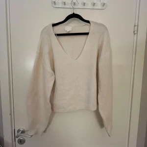 Vit ribbad stickad tröja med v-ringning - Mysig vit stickad tröja med djup v-ringning och ribbad struktur. Tröjan har långa ärmar och en något lös passform, perfekt för lager-på-lager-stil.