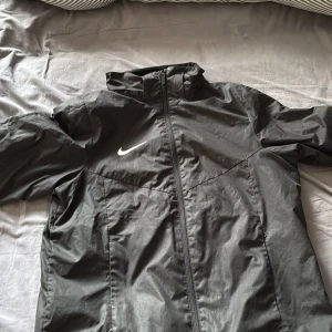 Svart vindjacka från Nike - Säljer en svart vindjacka från Nike med dragkedja framtill och hög krage. Jackan har en enkel och sportig design med Nike-logga på bröstet. Perfekt för utomhusaktiviteter när det blåser.