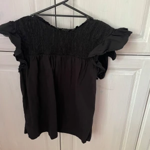 Svart blus  - Säljer för att den inte kommer till användning!!Säljer en svart blus från MNG med smock upptill, volangdetaljer på ärmarna och knytning i nacken. Blusen har korta ärmar och en luftig passform, perfekt för en trendig look. Skriv vid frågor 🥰