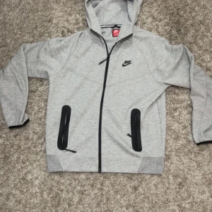 Grå Nike tech fleece (nya) - Säljer en helt ny oanvänd Nike tech fleece den nya versionen då den är lite för stor på mig. Den passar dig som har storlek M på tröjor! Jag har inga husdjur och röker ej så rökfri lukt och pälsfritt! Modellen är 178 cm lång och 60 kg
