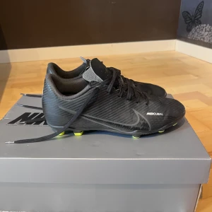 Nike Mercurial junior  - Svarta Nike Mercurial fotbollsskor med diskret randigt mönster och vita logotyper. Skorna har snörning och platt sula med gula dobbar för bra grepp på planen. Snygg, sportig design i syntetmaterial som ger en tight och smidig känsla när du spelar. Använda 6-7 gånger. 
