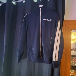 Palm Angels tech trackpants navy/blå och Palm angels tröja navy blå storlek M - Säljer ett par sjukt snygga tech trackpants från Palm Angels i marinblått med vita sidoränder och broderad logga på benet. Byxorna har elastisk midja och är gjorda i ett glansigt syntetmaterial som ger en sportig vibe. Perfekta för en streetwear-look.