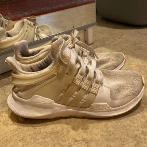 Beige Adidas sneakers - Säljer ett par vita sneakers från Adidas med klassiska tre ränder på sidan. Skorna har snörning och en platt sula. Ovandelen är i textil och syntetmaterial, med rund tå och bekväm passform. Dom är bara lite smutsiga men skulle tro att dom blir rena när man har    tvätttat dom 