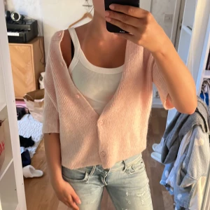 Ljusrosa stickad kofta - Säljer en ljusrosa stickad kofta med v-ringning och knappar framtill. Koftan är croppad och har en lös passform med kortare ärmar. Perfekt att slänga över ett linne eller t-shirt för en avslappnad look.