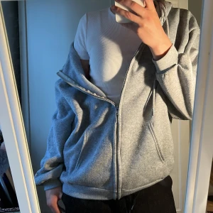 Grå zip-hoodie med huva - En klassisk grå baggy hoodie med dragkedja och huva. Tröjan har ribbade muddar, fickor framtill och justerbara snören i huvan. Perfekt för lager-på-lager och chill dagar. 