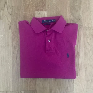 Rosa pikétröja från Polo Ralph Lauren - Snygg rosa pikétröja från Polo Ralph Lauren med klassisk krage och två knappar. Tröjan har korta ärmar och den ikoniska broderade loggan i mörkblått på bröstet. Tillverkad i mjuk bomull som känns skön mot huden. Väldigt skönt material samt matchar bra till ett par shorts inför sommaren! 