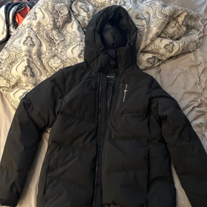 Svart pufferjacka från SAIL RACING - Svart pufferjacka från SAIL RACING med huva och dragkedja framtill. Jackan har en clean look med diskret logga på bröstet och bak på huvan. Perfekt för kalla dagar med sin vadderade design och långa ärmar. Snygg och enkel att matcha till allt.oanvänd köpt för 6999