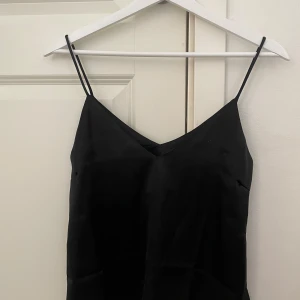 Svart silkestopp med smala axelband - En stilren svart linnetopp med tunna axelband och v-ringning. Toppen har en enkel och elegant design som passar perfekt till jeans eller kjol. Materialet känns mjukt och har en lätt glansig yta.