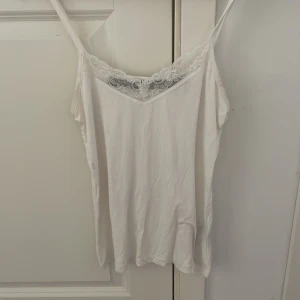 Vit linnetopp med spetskant - Säljer en vit linnetopp med tunna axelband och snygg spetsdetalj längs urringningen. Perfekt att styla lager på lager eller bära som den är under varma dagar. Enkel och stilren design som passar till mycket.