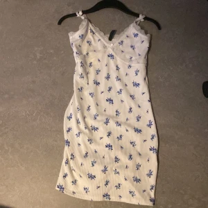 Vit blommig kortklänning H&M Divided - Supersöt vit kortklänning från H&M Divided med blått blommönster och spetsdetaljer vid v-ringad hals🤗Säljer då den aldrig kommit till användning.