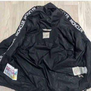 Svart vindjacka från Moncler med huva - Svart vindjacka från Moncler med huva och dragkedja framtill. Jackan har två snedställda fickor med dragkedja, Moncler-logga på ärmen och tryckt Moncler-text längs insidan av huvan. Lätt och smidig modell i syntetmaterial, perfekt för blåsiga dagar.
