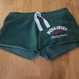 Gröna shorts från Hollister - Mörkgröna shorts från Hollister med vit snörning i midjan och broderad logga på ena sidan. Mjukt material och kort modell, perfekt för varma dagar eller avslappnade tillfällen.