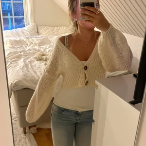 Beige stickad kofta med knappar - Säljer en beige stickad kofta med två knappar framtill. Koftan har en croppad passform, ballongärmar och är väldigt mjuk och mysig. Perfekt till sommarkvällar! 