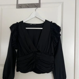 Svart croppad blus från Zara - Säljer en svart croppad blus från Zara med v-ringning, långa ärmar och volangdetaljer på axlarna. Toppen har snygg rynkning framtill och är tillverkad i ett lätt, glansigt material som ger en lyxig känsla. Perfekt för dig som gillar trendiga och feminina plagg.