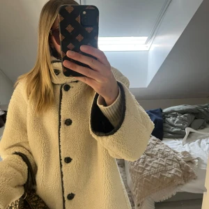 Vändbar teddyjacka i beige och brun - Säljer en vändbar teddyjacka med ena sidan i fluffig beige teddy och andra sidan i mörkbrun mockaliknande material. Jackan har stora svarta knappar, bred krage och långa ärmar. Håller värmen jättebra