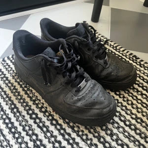 Svarta Nike Air Force 1 sneakers - Klassiska helsvarta Nike Air Force 1 sneakers i skinn med lågt skaft och snörning. Snygg och tidlös modell med rund tå och perforerade detaljer på tån för extra ventilation. Perfekta för dig som gillar streetstyle och vill ha en ikonisk sneaker i garderoben. 🚨Inga inre sulor🚨