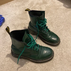 Gröna Dr. Martens boots i skinn - Snygga gröna Dr. Martens boots i glansigt skinn med klassisk gul söm runt sulan. Höga skaft, rund tå och gröna snören. Perfekta för dig som vill sticka ut med en cool färg på dina boots. Synligt använda med lite repor och creases (syns på bild).