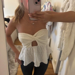 Vit bandeau tubtopp med broderad volang - Supersöt vit bandeau tubtopp med twistad front och liten cut-out. Överdelen är virkad och nederdelen har broderad volang med hålmönster. Perfekt till sommaren och ger en romantisk vibe. Sitter snyggt och ärmlös för en luftig känsla.