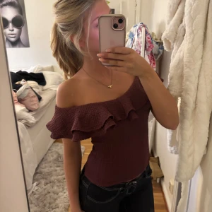 Vinröd offshoulder topp med volang - Säljer en vinröd offshoulder topp med bred volang runt hela axelpartiet. Toppen är figurnära och har en smockad struktur som ger en snygg passform. Perfekt att styla med jeans eller kjol för en trendig look.