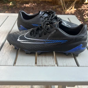Nike phantom gx academy  - Säljer ett par svarta Nike phantom academy fotbollsskor från Nike med blå detaljer och vit swoosh. Skorna är i bra condition och har använt dom fåtals gånger. Skorna är size 44,5 men passar 43. Perfekta för dig som vill sticka ut på planen med stilrena och sportiga skor.