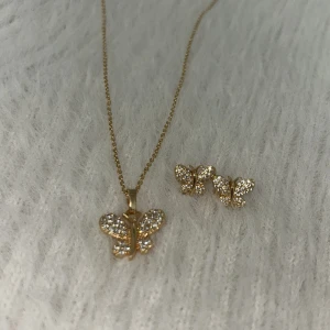 Glittrigt fjärilssmyckeset i guld - Superfint smyckeset med halsband och örhängen i form av fjärilar. Både halsbandet och örhängena är i guldton och dekorerade med glittrande stenar som ger en lyxig känsla. Perfekt för dig som älskar detaljer och vill sticka ut med något extra sött.