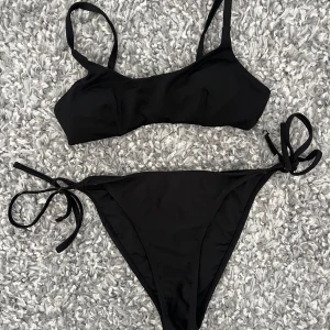 Svart bikini med knyt i sidorna - Stilren svart bikini med enkel topp och klassiska bikinitrosor med knyt i sidorna. Bikini från NAKD. Båda delarna Xs