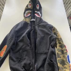 Svart och camo hoodie från A Bathing Ape - Säljer en svart hoodie från A Bathing Ape med ikonisk hajdesign på huvan och camo-detaljer på ena ärmen och huvan. Dragkedja framtill och broderade detaljer på ärmarna. Perfekt statement-plagg för streetwear-stilen. Dragkedjan är lite trög men den funkar. Går ner i pris vid snabb affär!