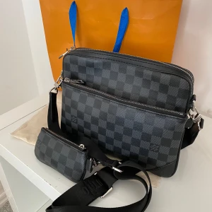 Svart axelväska från Louis Vuitton med rutigt mönster - Snygg svart axelväska från Louis Vuitton med klassiskt rutigt mönster. Väskan har flera fack med dragkedja och en bred justerbar axelrem med logotyp. Tillverkad i syntetmaterial och har en mindre avtagbar väska som detalj.