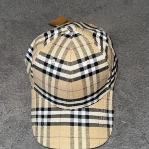 Burberry keps - Säljer en klassisk rutig keps från Burberry i beige, svart och vit. Kepsen har det ikoniska rutmönstret och justerbar rem baktill. Perfekt accessoar för en stilren look.