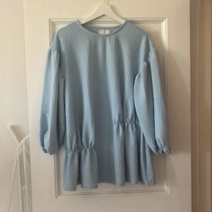 Ljusblå långärmad klänning med volang - Supersöt ljusblå klänning med lång ärm och volangdetaljer vid midjan och nederkanten. Klänningen har en lös passform och är tillverkad i ett strukturerat tyg som ger en extra touch. Perfekt för vår och sommar!