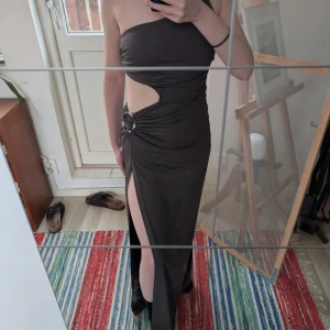 Mörkgrå långklänning med slits och cut-out - Säljer en snygg mörkgrå långklänning med en axel, cut-out vid midjan och hög slits framtill. Klänningen har en dekorativ ringdetalj i sidan och är figurnära. Perfekt för fest eller bal! Materialet är mycket stretchigt och följsam!.