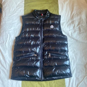 Svart dunväst från Moncler - Snygg svart dunväst från Moncler med glansig finish och klassisk quiltad design. Västen har hög krage, två dragkedjefickor framtill och Moncler-logga på bröstet. Perfekt för lager-på-lager och till sommaren.