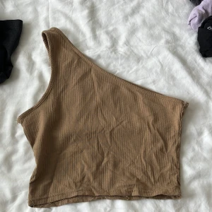 Beige ribbad oneshoulder topp - Snygg beige ribbad topp med oneshoulder-design. Perfekt för dig som vill ha en enkel men trendig look. Toppens ribbade material ger en cool struktur och sitter skönt mot kroppen.